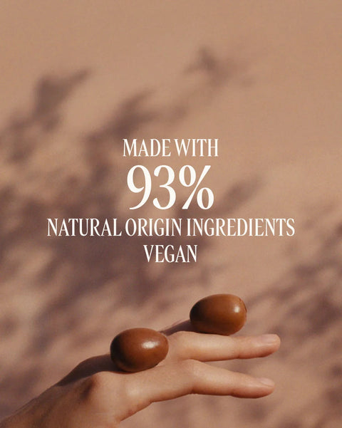 A hand holding two smooth brown oval objects against a warm beige background with soft shadows, overlaid with the text 'MADE WITH 93% NATURAL ORIGIN INGREDIENTS VEGAN'.