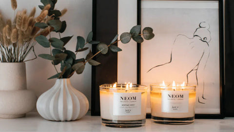 Two NEOM candles on a surface with decorative elements including a vase with plants and a framed artwork.