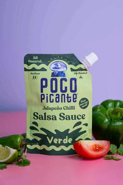 A stand-up squeeze pouch of POCO Picante Jalapeño Chilli Salsa Sauce in Verde flavor, styled on a pink surface with green bell peppers, lime, and tomato slices against a lavender background.