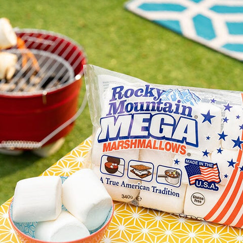 A bag of Rocky Mountain Mega Marshmallows placed on a yellow patterned blanket outdoors, with a small bowl of white marshmallows in the foreground and a red grill blurred in the background on green grass.