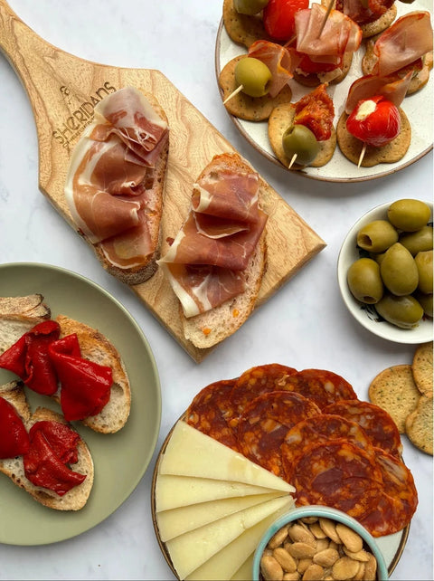 An overhead styled flat-lay of charcuterie boards and appetizers on a white marble surface. Visible items include sliced bread topped with prosciutto, red sunburst tomatoes, olives on skewers, crackers, sliced cheese, salami, almonds, and a wooden serving board engraved with the brand name 'Shampions'.