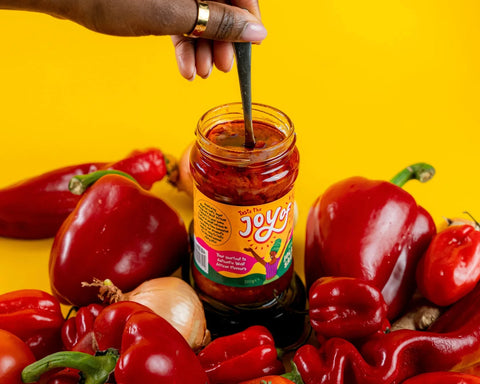 A glass jar of Joy of the Pantry hot sauce is shown with a hand holding a spoon above it, surrounded by fresh red bell peppers and a whole onion on a bright yellow background.