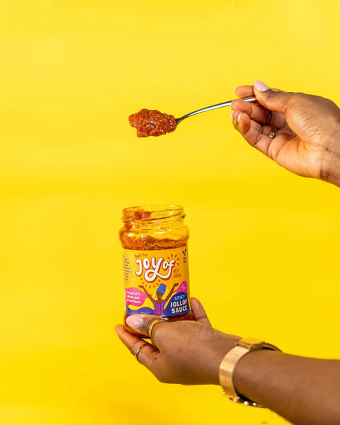 A hand holds a spoon with red chili sauce above a glass jar of Joy of India's Hot Habanero Chili Sauce against a bright yellow background.