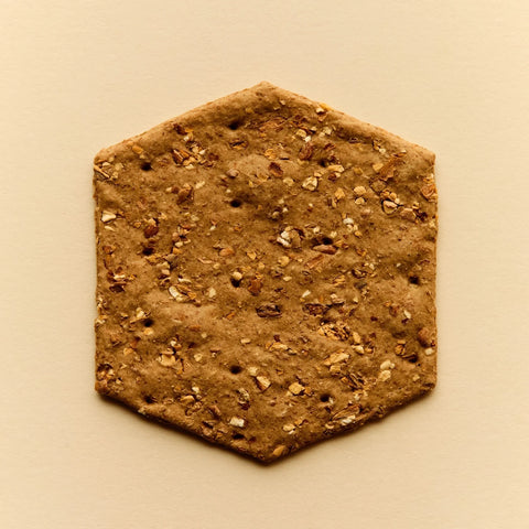 A hexagonal flatbread or cracker cookie with visible oats and nuts, photographed from directly above on a solid beige background.