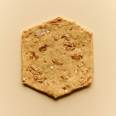 Hexagonal flatbread or cracker with embedded oats and small white flakes, photographed from above on a warm beige background.