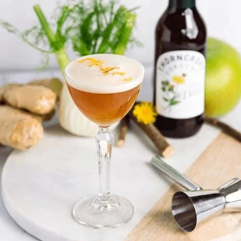 A frothy amber cocktail in a coupe glass, styled on a white board with ginger roots, a dark bottle, a jigger, and fresh herbs in the background.