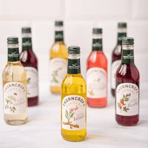 A group of Thorncroft Organic ginger beer bottles arranged on a light surface, with one bottle in the foreground labeled Ginger and several other flavor bottles blurred in the background.