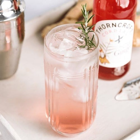 A pink cocktail in a clear ribbed glass with ice, garnished with a sprig of rosemary, set on a light surface with a bottle of Thorncroft Cordial Ginger behind it.