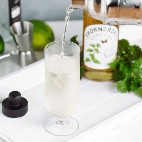 A clear glass cocktail glass with a stem, partially filled with a pale frothy drink, shown on a white tray with a bottle of Thorncroft Cordial, a jigger, lime, and parsley in the background.