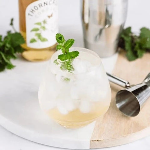 A frothy pale yellow cocktail in a clear stemless glass with ice, topped with a mint sprig, styled on a white marble board with a jigger, metal shaker, and a bottle in the background.
