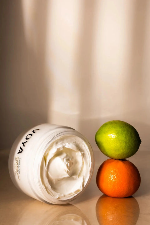 An open white jar of Voya cream or balm lies on its side on a reflective beige surface, with a green lime and an orange stacked beside it. The background is a softly lit cream curtain with warm shadows.