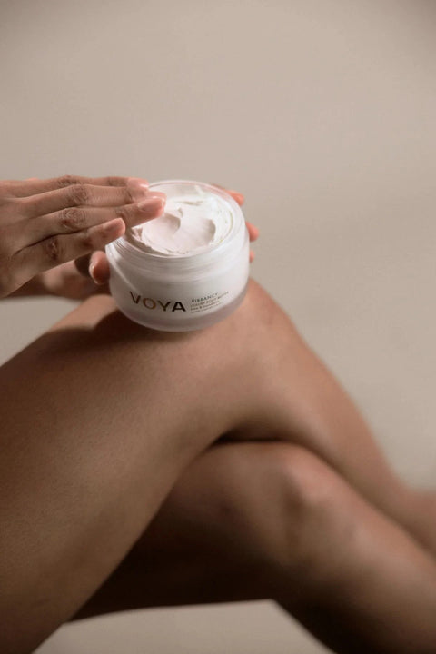 A person holding an open jar of VOYA skincare cream against their leg, showing the white cream inside. The background is a soft, neutral beige studio setting.