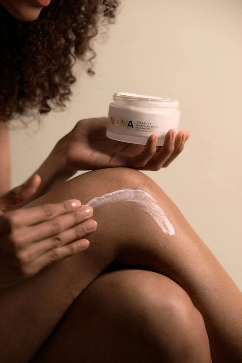 A person applies a white cream to their knee while holding a white jar of VONA Radiance Body Butter against a warm beige background.