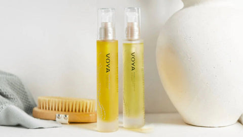 Two bottles of Voya product on a white surface with a brush and towel in the background.
