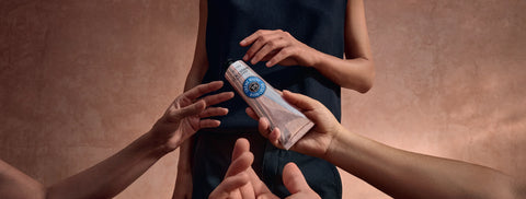 A hand cream tube held by hands in front of a person wearing a dark sleeveless top, set against a warm textured brown background.