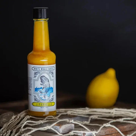 A bottle of White Whale Sauce Ginger Bender hot sauce with a black cap, standing on a rustic wooden surface with a net and shell in the foreground and a blurred lemon in the background against a dark backdrop.