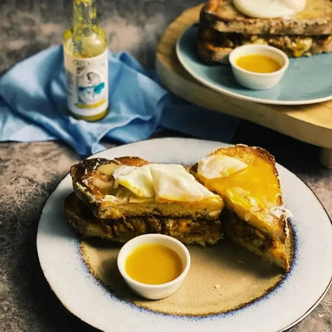 A plated breakfast food scene showing two halves of a toasted sandwich with a poached egg and yellow sauce, served on a ceramic plate with a small sauce dish. A second plate and sandwich are blurred in the background, along with a bottle and blue cloth on a tabletop.