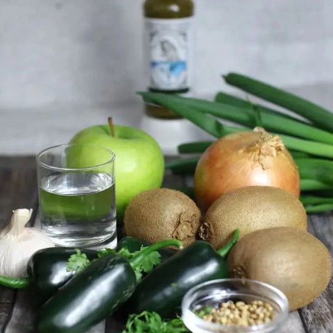 A styled flat-lay of fresh ingredients on a rustic wooden surface, including a green apple, an onion, several kiwis, jalapeños, garlic, scallions, cilantro, a glass of water, and a small bowl of seeds, with a bottle blurred in the background.